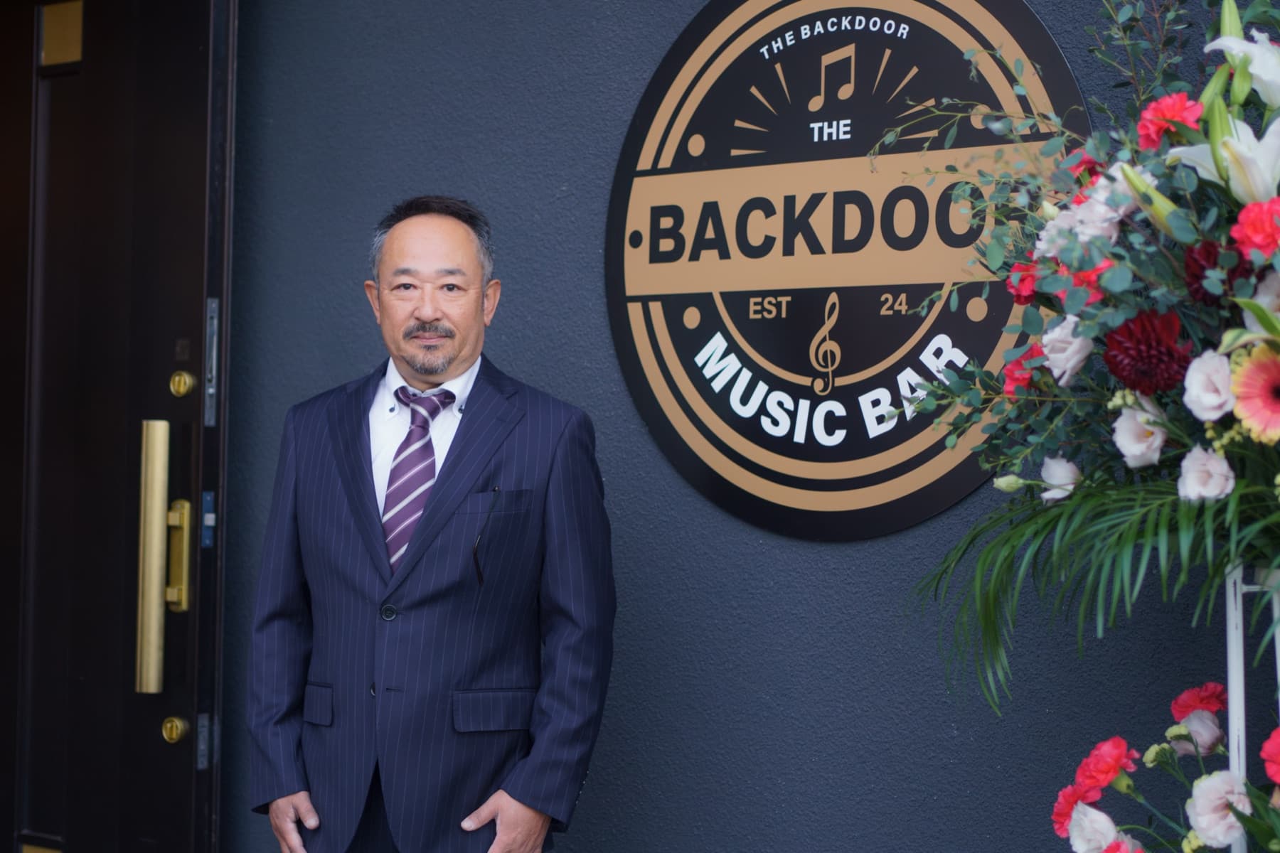 Entrance to The Backdoor — the owner standing beside the large brass Backdoor badge mounted on the exterior wall, a floral arrangement to the right.