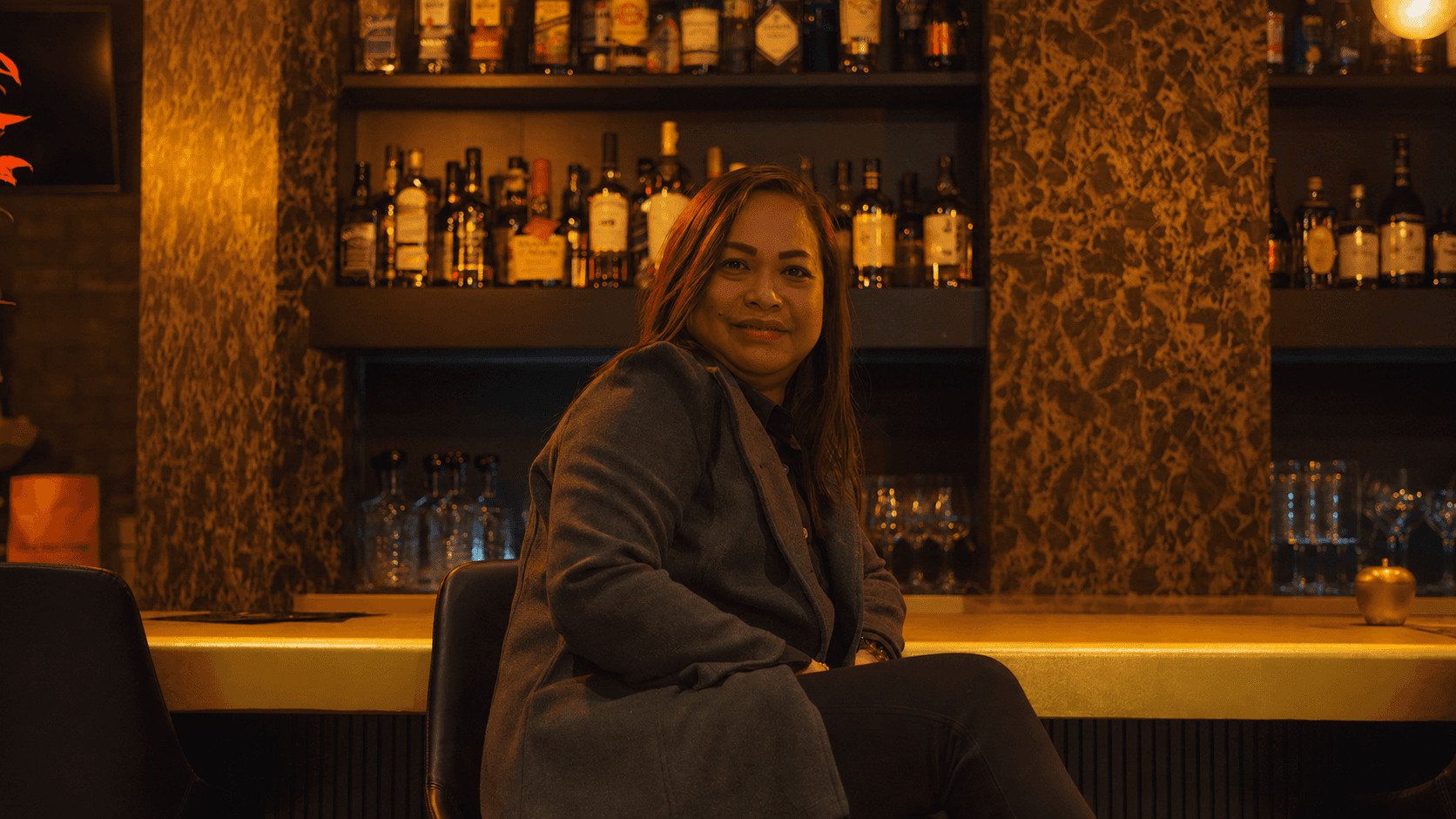 Manager seated at the bar counter. Long hair, dark coat, warm light from the whisky shelf behind.